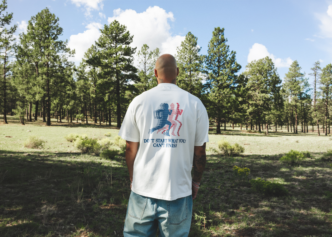 Model seen from behind wearing a Compliments white t-shirt with a 'Don't start what you can't finish' back print, standing in a sunlit pine field.